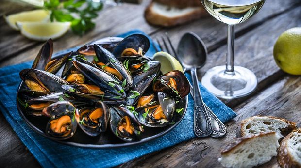High angle view of steamed mussels in black plate and white wine glass shot on wooden table.  Sliced ​​lemon and bread round out the composition.  Choice of focus on mussels.  The dominant colors are black, orange and blue.  XXXL 42Mp studio photo taken with Sony A7rii and Sony FE 90mm f2.8 macro G OSS lens