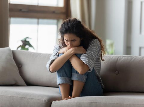 Stressed young woman embracing knees, sitting alone on couch in living room, feeling depressed. Unhappy desperate millennial curly lady regretting of decision, thinking of personal problems at home.