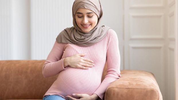 Cheerful Pregnant Muslim Lady In Hijab Posing Embracing Belly Sitting On Couch At Home. Young Arab Mom's Portrait, Awaiting Baby. Pregnancy Happiness And Childbirth Concept