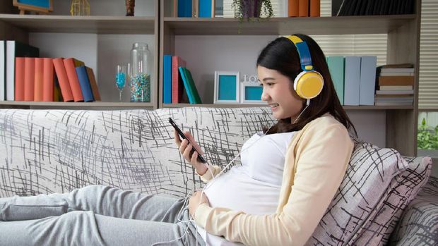 Beautiful young pregnant woman listening to music and sitting on sofa at living room. Relax, rest time concept.