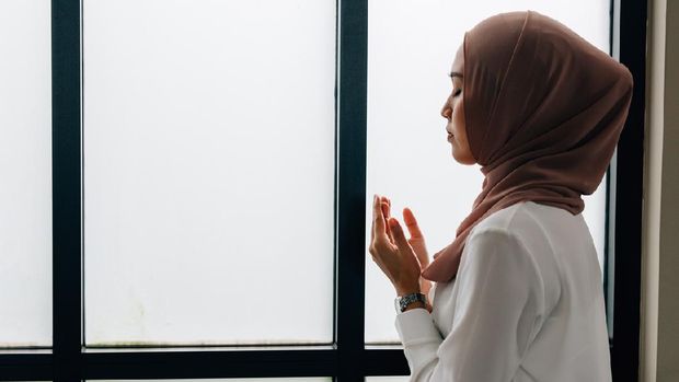 Southeast Asian Islamic female in hijab clasping hands and looking up while standing near window during prayer