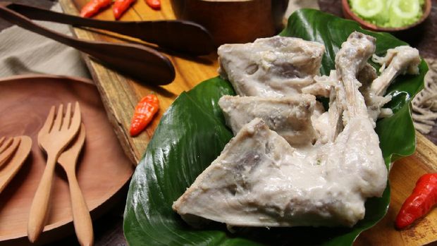 Ayam Pop, a popular chicken dish from Minangkabau, Padang, West Sumatra. The chicken is braised in coconut milk and spices; then only lightly fried. It is served with a special spicy paste of red chili peppers, tomatoes, and broad beans. The chicken pieces are arranged on rectangular wooden serving block that has been lined with a broad green leaf. On the other end is the chili paste. Some chili peppers are scattered on the tray. Some green vegetables of cassava leaves, lettuce and cucumber slices are also served along the chicken dish. A wooden plate with two matching wooden cutleries are placed next to the tray.