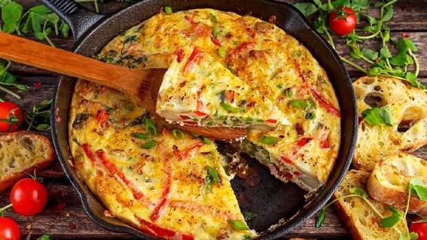 Frittata made of eggs, potato, bacon, paprika, parsley, green peas, onion, cheese in iron pan on wooden table