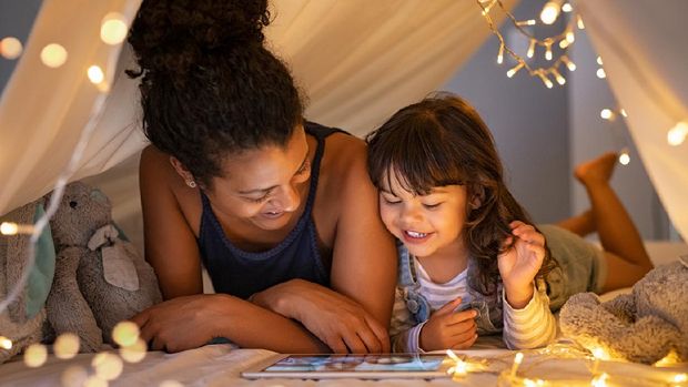 African mother and cute smiling girl using digital tablet while lying in illuminted tent in kid bedroom. Cheerful ethnic woman and lovely daughter on video call under a cozy hut. Lovely little girl with mom watching cartoon on digital tablet in bedroom.