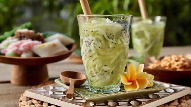 Ilustrasi es buah selasih Es Kuwut, a Balinese cold dessert drink of honeydew melon, coconut, basil seeds, and lime. The drinks are served in clear tall drinking glasses and each garnished with lime half, lime leaves, and a bamboo straw. Each glass is placed on a ceramic saucer embossed with frangipani flower. Fresh frangipani flowers and a wooden spoon are also placed on the saucer. The saucer is then arranged on large wooden coaster carved with flowery shape; together with local snacks of traditional sweet cakes and roasted peanuts. The image was shot outdoor on the terrace with natural lighting. The green leaves on the trees in the garden become the blurry background.