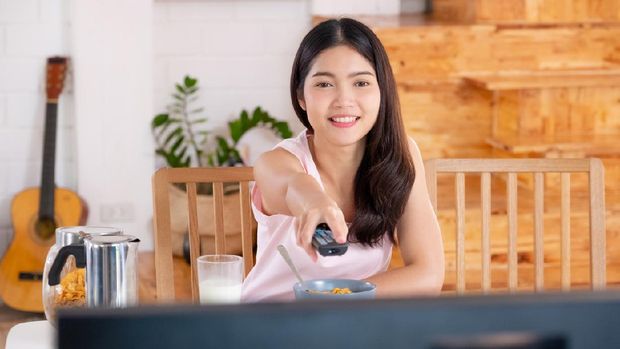 asian woman wathcing tv on demand and use remote control change television channel at breakfast in morning