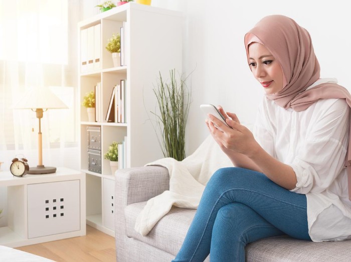 1015858366 elegant attractive female muslim using mobile ponsel pandai searching online information when she sitting on sofa couch relaxing.