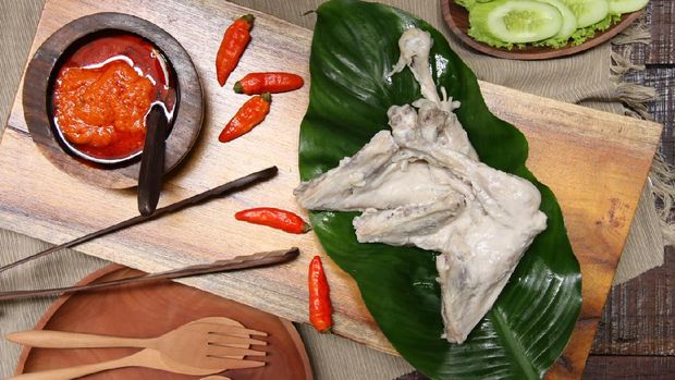 Ayam Pop, a popular chicken dish from Minangkabau, Padang, West Sumatra. The chicken is braised in coconut milk and spices; then only lightly fried. It is served with a special spicy paste of red chili peppers, tomatoes, and broad beans. The chicken pieces are arranged on rectangular wooden serving block that has been lined with a broad green leaf. On the other end is the chili paste. Some chili peppers are scattered on the tray. Some green vegetables of cassava leaves, lettuce and cucumber slices are also served along the chicken dish. A wooden plate with two matching wooden cutleries are placed next to the tray.