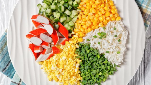 Bahan sup jagung kepiting Crab salad of surimi sticks with canned corn, cucumbers, spring onion hard-boiled eggs, jasmine rice divided into sectors on a white plate on a white wooden background, top view, close-up