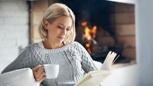 A beautiful young woman reading a book and enjoying a warm beverage near a fireplace