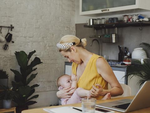 A portrait of a beautiful young Caucasian mother breastfeeding her adorable baby girl while working on a laptop in a kitchen at home.