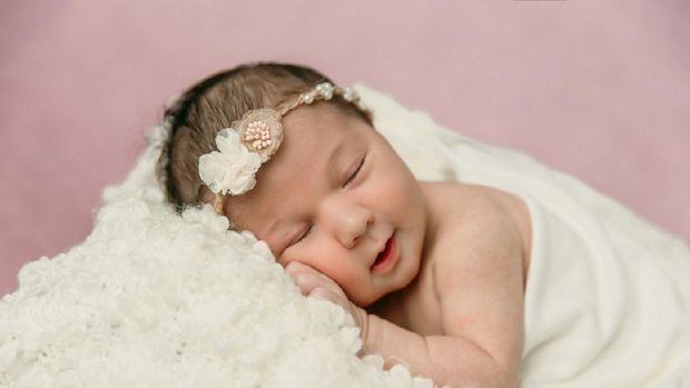 Sleeping newborn baby girl laying on a cream colored blanket on a pink purple backdrop with a headband in her hair and a slight smile