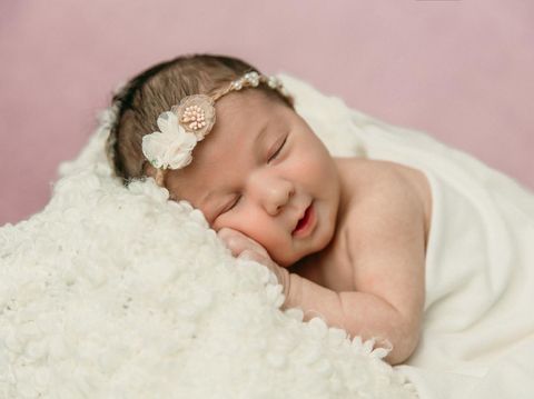 Sleeping newborn baby girl laying on a cream colored blanket on a pink purple backdrop with a headband in her hair and a slight smile