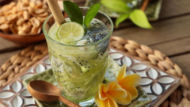 Es Kuwut, a Balinese cold dessert drink of honeydew melon, coconut, basil seeds, and lime. The drink is served in a clear tall glass and garnished with lime half, lime leaves, and a bamboo straw. The glass is placed on a ceramic saucer embossed with frangipani flower. Fresh frangipani flowers and a wooden spoon are also placed on the saucer. The saucer is then arranged on large wooden coaster carved with flowery shape; together with local snack of roasted peanuts. The image was shot outdoor on the terrace with natural lighting.