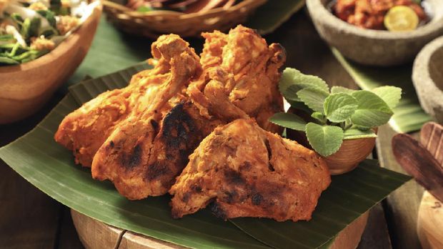 Ayam Bakar Padang, the traditional spicy grilled chicken from Minangkabau cuisine, West Sumatra. The chicken pieces are arranged on a wooden cutting block lined with banana leaf. Accompanied with red chili sauce and a vegetable salad.