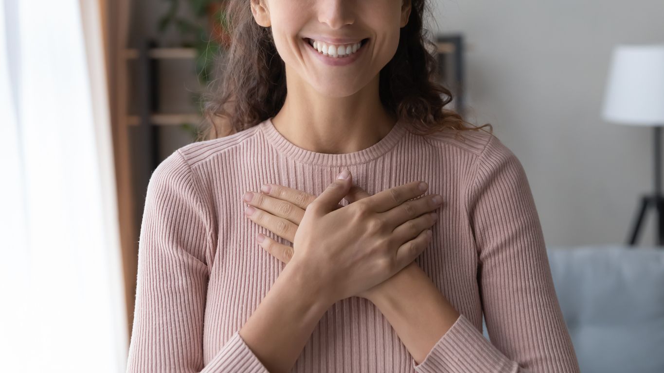 Close up focus on happy sincere female holding folded hands on chest. Emotional positive kind candid millennial woman feeling thankful indoors, showing gratitude sign, believe faith charity concept.