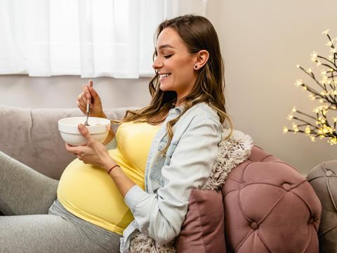 Beautiful young pregnant woman relaxing on sofa and eating corn flakes. Pregnancy concept.