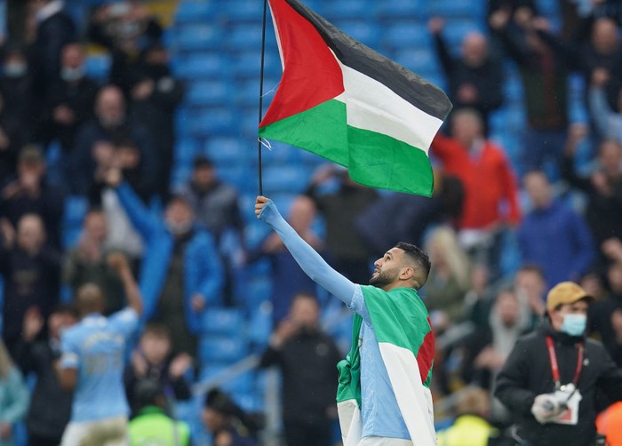 MANCHESTER, ENGLAND - MAY 23: Riyad Mahrez of Manchester City wears the flag of Algeria, as he carries the flag of Palestine after Manchester City are presented with the Premier League Trophy following their victory in the Premier League match between Manchester City and Everton at Etihad Stadium on May 23, 2021 in Manchester, England. A limited number of fans will be allowed into Premier League stadiums as Coronavirus restrictions begin to ease in the UK. (Photo by Dave Thompson - Pool/Getty Images)