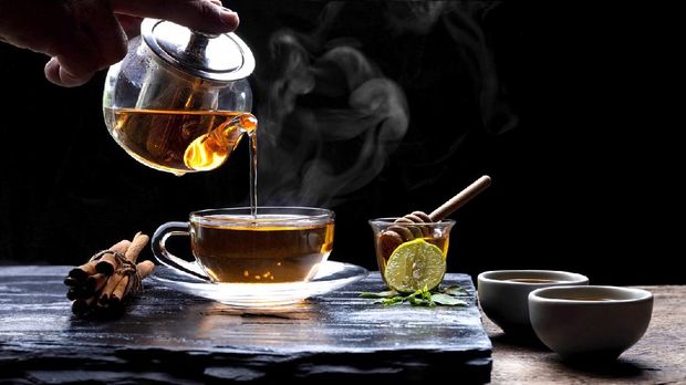 Pouring hot aromatic herbal tea from teapot into glass teacup set with steam and various herbs on black stone plate with wooden table floor in dark background