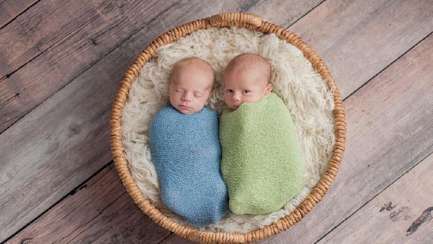 Four week old fraternal, twin baby boys swaddled in light blue and green wraps and lying in a wicker basket. One brother appears to be whispering a secret to the other brother.
