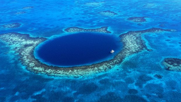 An aerial view of a boat moored in the Great Blue Hole off the coast of Belize. The azure seas of the Caribbean envelope the deep blue of the Great Blue Hole. A coral reef surrounds the geologic wonder.