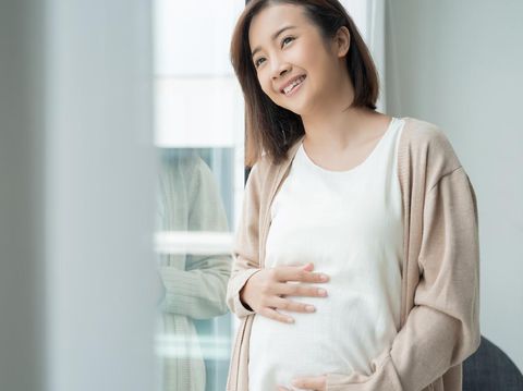 Young Asian pregnancy woman holding tummy with both hands sitting on sofa at home. Pregnant girl look, touch at stomach. Feeling of baby inside belly. Thinking of newborn infant be delivered in future