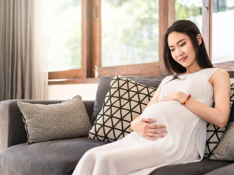 Young Asian pregnancy woman holding tummy with both hands sitting on sofa at home. Pregnant girl look, touch at stomach. Feeling of baby inside belly. Thinking of newborn infant be delivered in future
