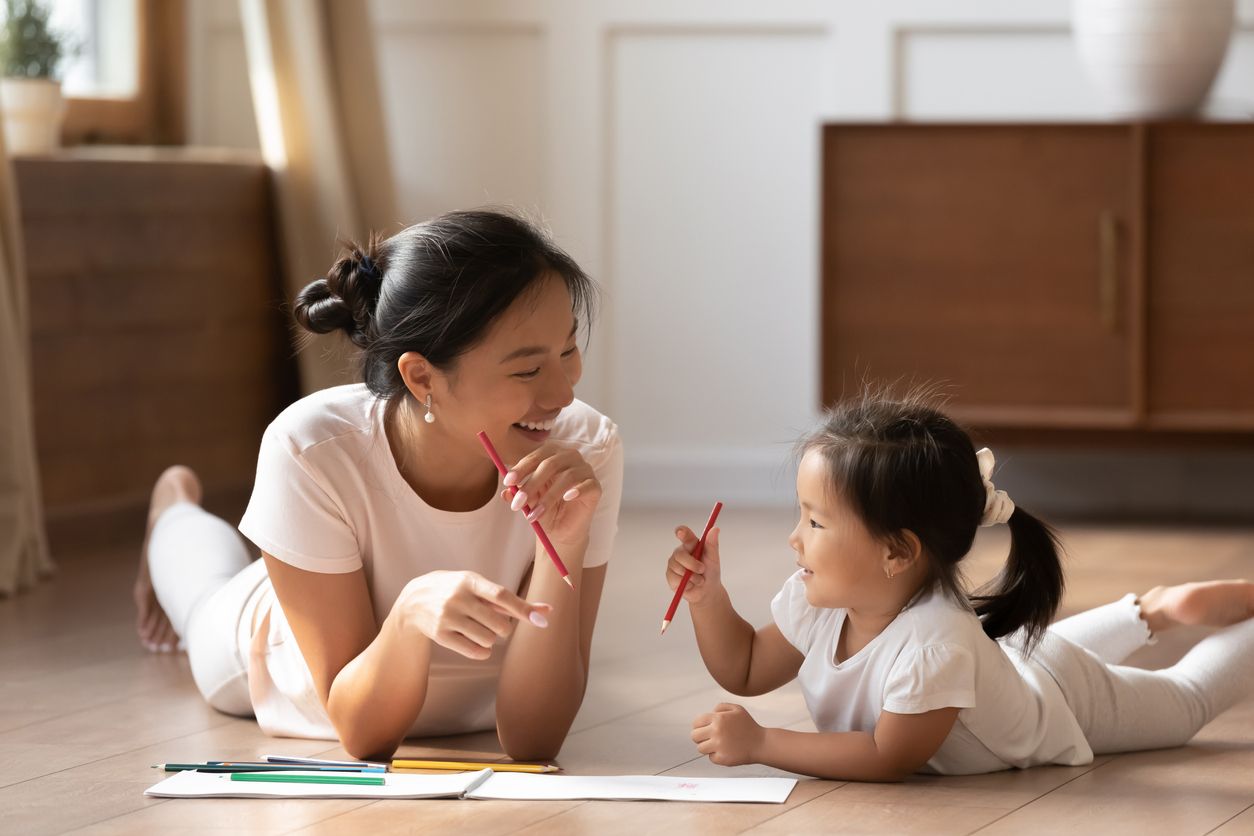 Educational pastime develop creativity skill in kid concept. Asian mother her small daughter lying on warm wooden floor in sunny cozy living room, mom teach girl paint use album and colourful pencils