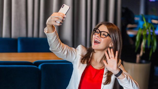 Portrait of happy joyful beautiful stylish brunette young woman in glasses sitting on video call or doing selfie with toothy smile and happiness waving hand. indoor studio shot, cafe office background