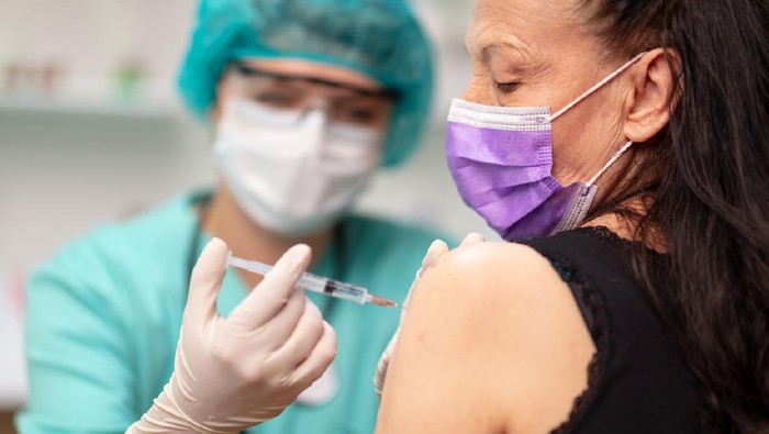 Senior woman wearing protective face mask getting flu shot, female doctor in protective workwear holding syringe and injecting vaccine in patients arm; prevention and immunization from corona virus infection