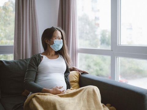 worried pregnant woman sitting at home with protective face mask and looking through the window.
