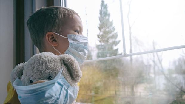 Sad illness child on home quarantine. Boy and his teddy bear both in protective medical masks sits on windowsill and looks out window. Virus protection, coronavirus pandemic, prevention epidemic.