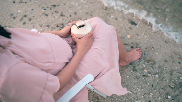 Cheerful pregnant woman having fresh coconut on the beach sitting on a picnic foldable chair by the seaside and enjoying the sunset. Summer vacation during pregnancy and outdoors lifestyle