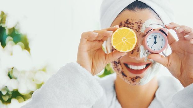 Young woman in bathrobe and wrapped in towel holding a lemon and clock over her eyes. It's always time to take care of your skin.