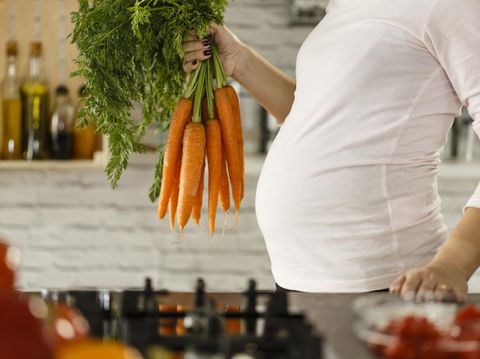 Pregnant mid adult woman holding bunch of carrots in the kitchen, preparing food.