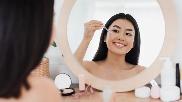 Eye zone care concept. Happy beautiful asian girl applying eye serum in front of mirror at home