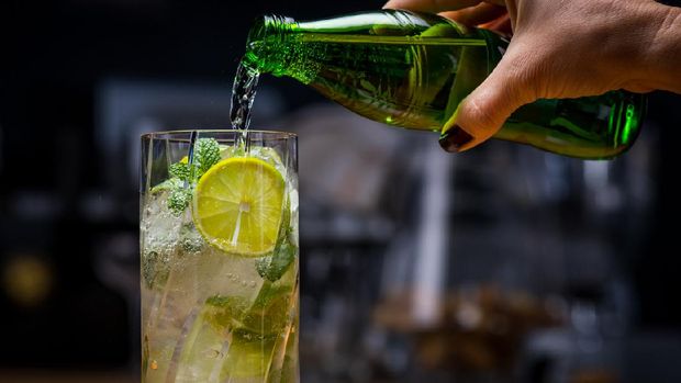 Close-up of female bartender adding sparkling water to cocktail with lemon and mint leaves.