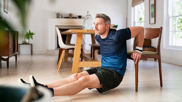 Mature man doing fitness exercise while doing pushups using chair in living room. Middle aged man training at home wearing sportswear during fitness exercises for triceps. Mid adult fit guy exercising for wellness in his living room.