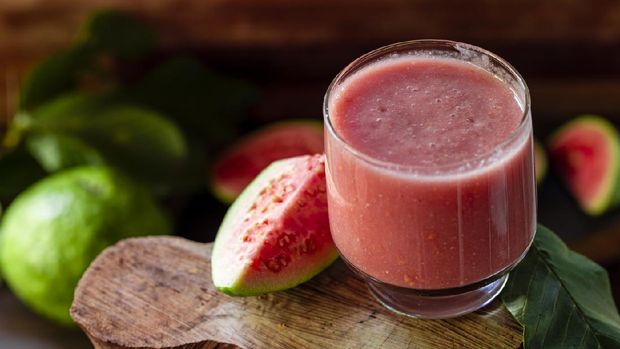 Delicious red guava juice alongside a slice of guava, in the background guavas and leaves on rustic wood