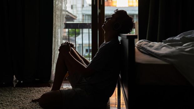 Wide shot young adult asian loneliness sad man sitting on the floor in bedroom leaning on bed and cry. Broken heart despair man sitting on the floor listening music from headphone alone.