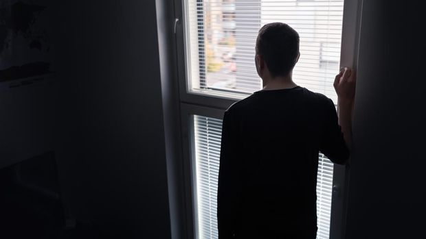 Lonely man looking out the window. Solitude, isolation and loneliness concept. Sad upset person with regret, shame or stress in moody dramatic back view silhouette. Dark room with shadow at home.