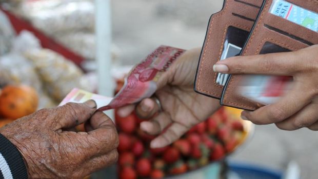 Seorang wanita muda mengambil uang rupiah dari dompetnya untuk membayar beberapa barang kepada wanita tua di pasar lokal Bali.  Transaksi kecil.  Selamat berbelanja.