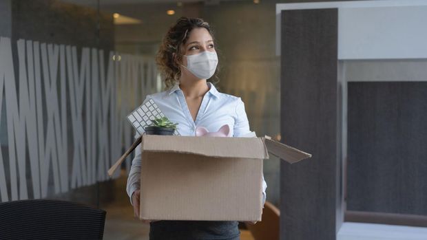 Female worker wearing a facemask at the office while holding a box with her belongings after being fired from her job during the COVID-19 pandemic