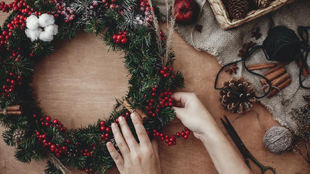 Rustic Christmas wreath flat lay. Hands holding fir branches, red berries and pine cones,thread, scissors, cinnamon, cotton on rustic wooden background. Making wreath at workshop