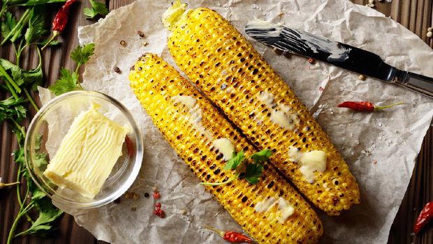 Top view of kitchen table with grilled sweet corn cob under melting butter and greens on baking paper