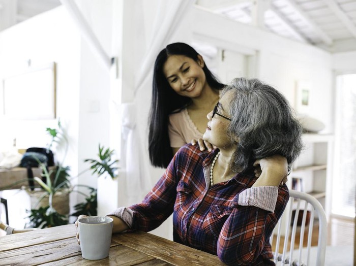 Beautiful adult granddaughter hugging beautiful senior grandmother while sitting on the chair drinking coffee