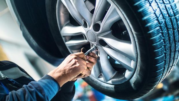 Closeup of unrecognizable male mechanic unscrewing the bolts on the rim of car wheel in order to replace tyre or the wheel itself. After he got those bolt loose with power tool he's using hand screwdriver to pull them out. The mechanic is wearing blue working uniform and the wheel is lit with blue fill light. The car is lifted up on a car jack