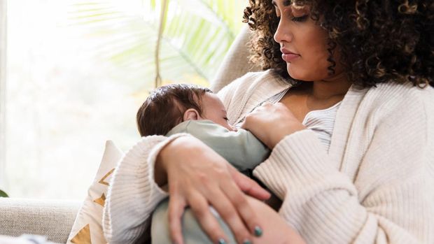 Sitting on a chair in the living room, the new mom watches as her baby boy breastfeeds.