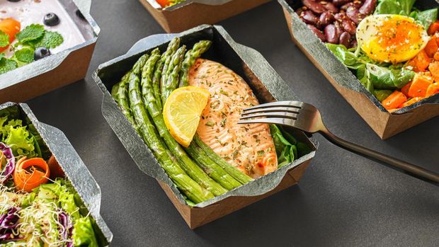 Ready healthy food catering menu in lunch boxes fish and vegetable packages as daily meal diet plan courier delivery with fork isolated on black table background. Take away containers order concept.