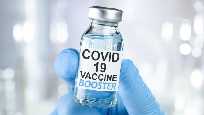 Hand in blue medical gloves holding a vaccine vial with Covid 19 Vaccine Booster text, for Coronavirus booster shot.